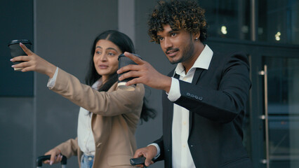 Talking business coworkers colleagues in city smiling Arabian man businessman Indian woman businesswoman arrive in city airport station with luggage baggage walking holding coffee pointing direction