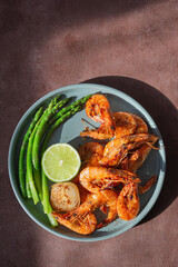 Grilled shrimp (tiger prawns) in spices with asparagus and a slice of lime (lemon) on a plate on a brown background.