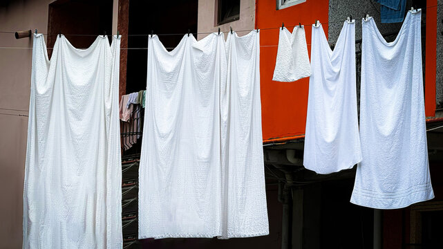 Freshly washed white sheets hanging on clothesline outside, swaying in breeze against building backdrop. Emphasizes simplicity and freshness of air-dried laundry and sustainable living practices