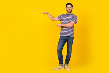 Full length photo of cheerful guy wear striped t-shirt jeans indicating at offer under arm empty space isolated on yellow color background