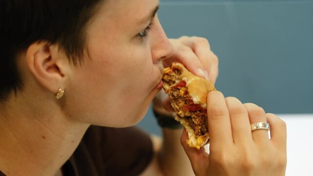 A young woman with stylish short haircut is sitting in a cafe and biting a delicious fresh hot meat burger with cheese. Girl in a cafe enjoying a break alone with cheeseburger. Close view