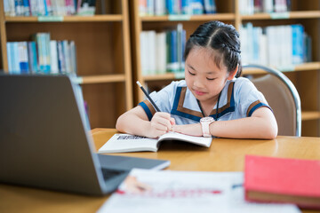 Young girl in a school uniform studying alone at a desk, writing in a notebook while referencing a laptop, smiling and focused, surrounded by bookshelves, capturing a moment of independent learning