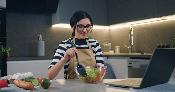 Smiling woman using laptop computer and mixing salad in glass bowl using spoon in modern kitchen
