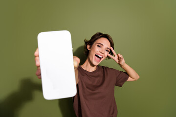 Photo portrait of pretty young girl hold telephone show v-sign excited dressed stylish brown outfit isolated on khaki color background