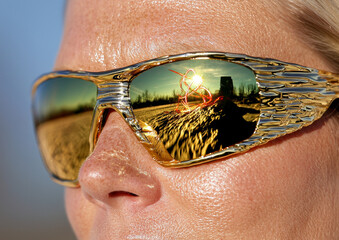 Close-up of a woman's face wearing gold mirrored sunglasses, reflecting the sun and a blurry cityscape.