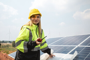 Smiling female engineer standing in front of solar panels looking at the camera and holding a walkie talkie to communicate with the team.