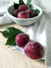 Fresh ripe plums in a ceramic bowl with leaves on a light wooden surface. Image for menu and cafe design