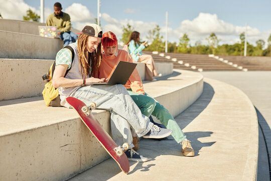 Teenage male skateboarder using laptop surfing net together with female friend sitting on concrete seat of amphitheater in park while studying outdoors enjoying sunny day, copy space