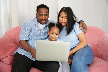 African American father and asian mother with daughter using laptop computer for watching movie or browsing internet on sofa