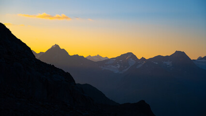 The first light of day casts a warm glow over the Otztal Alps, highlighting the rugged silhouette of majestic peaks against a vibrant sky filled with soft hues of orange and yellow.