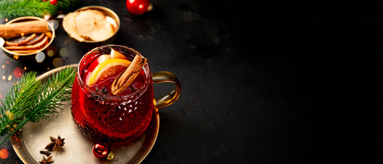 A glass mug of mulled wine on the black background and Christmas decorations. Festive winter drink, web banner with copy space for text
