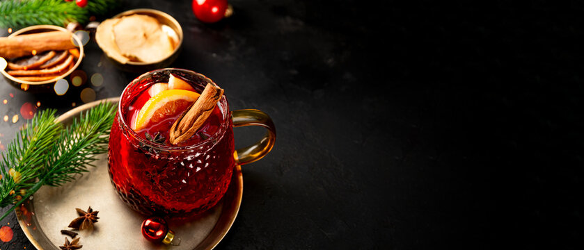 A glass mug of mulled wine on the black background and Christmas decorations. Festive winter drink, web banner with copy space for text