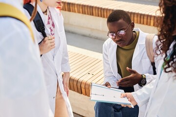 Medium shot of young male African American med student in white coat reviewing material in notebook discussing notes with classmates while studying outdoors on campus