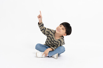 Happy Asian little boy pointing up with empty copy space and sitting on floor isolated on white...