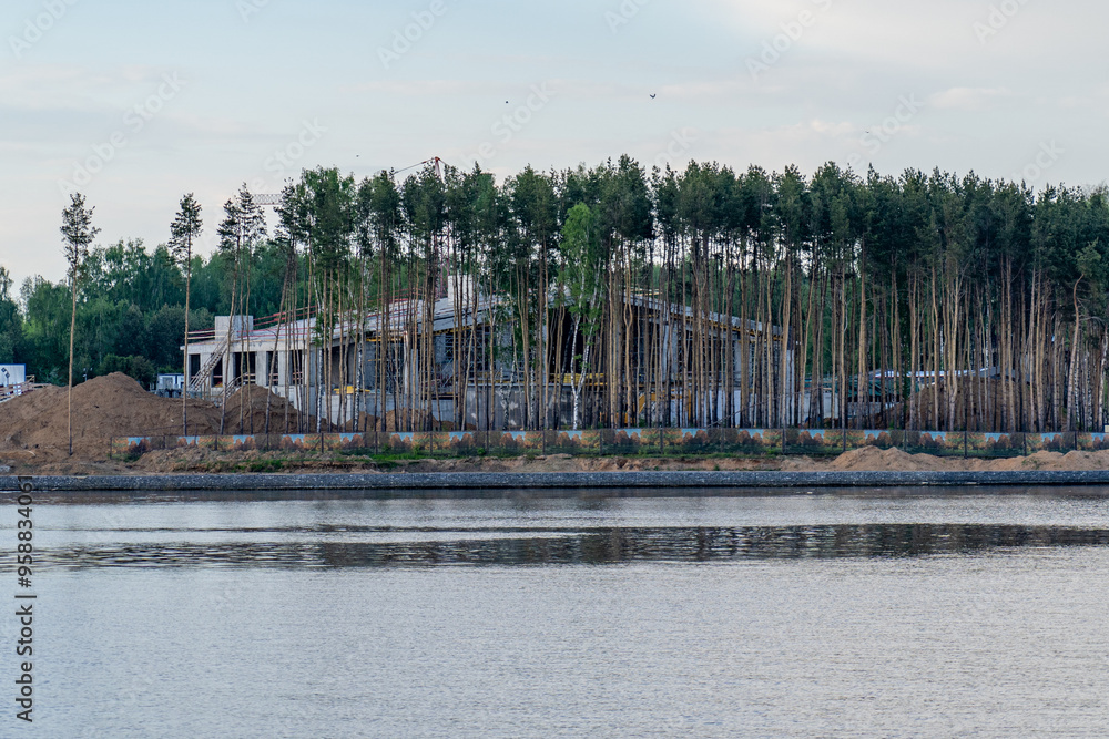 Poster a developing structure is being built beside a calm river, with trees lining the horizon under a cle