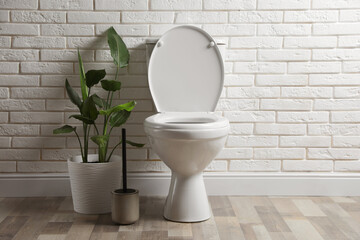 Ceramic toilet bowl, brush and houseplant near white brick wall in restroom
