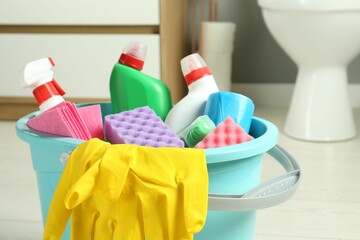 Bucket with different toilet cleaners, rag, sponge and gloves in bathroom, closeup