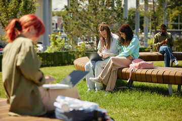 Full shot of two college students on bench using laptop together while studying outdoors on campus with green lawn enjoying fresh air and sunny day