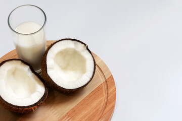 Coconut halves with pulp next to a glass of coconut milk on top of a round wooden board photographed from top to bottom