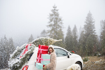 Woman holds a heap of gift boxes while standing by the car decorated with Christmas wreath and tree...