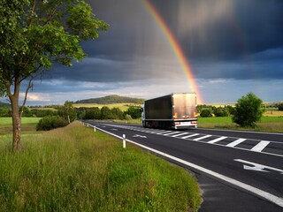 Motion blurred truck driving on asphalt road in rural landscape with colorful rainbow, rain and storm clouds