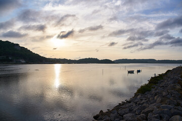 Sunset on a calm lake with the sun rising between the mountains with the sky covered by clouds and a small wooden boat in the background with its reflection in the water