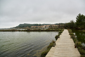 Wooden walkway coming out from the right of the image surrounded by water with a town on the horizon and the sky with grey clouds