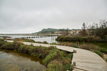 Wooden walkway with several small curves surrounded by water and vegetation with a town on the horizon with the sky with gray clouds