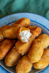 A vibrant blue plate filled with delicious, golden-brown croquettes resting on a soft green fabric background. One croquette is split open, revealing the creamy and rich bechamel filling inside, showc