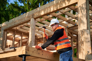 Carpenter constructing wooden frame house near forest. Bearded man with glasses hammering nails...