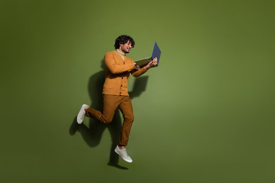 Full length photo of handsome young guy hold netbook running dressed stylish brown garment isolated on khaki color background