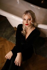 A glamorous blonde woman in a black velvet outfit sits elegantly by a vintage clawfoot bathtub in a...