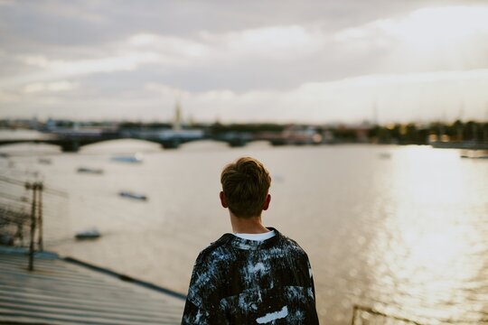 Individual gazing at city skyline from elevated platform near water. Sunlight softly illuminating the scene while boats dot the river in background