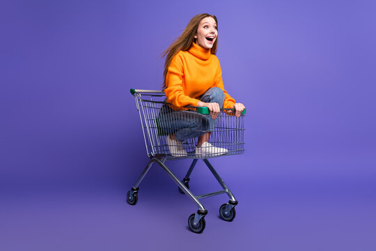 Photo of blonde youngster girl in orange turtleneck and jeans riding supermarket cart having fun adventures isolated on purple color background