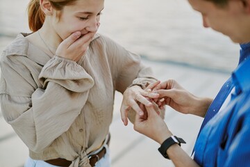 Woman having emotional response while receiving engagement ring from partner in waterfront setting,...
