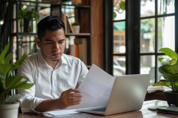 Young busy latin professional business man checking document working at laptop computer in office. Serious businessman accountant expert reading legal paper company file overview at workplace.