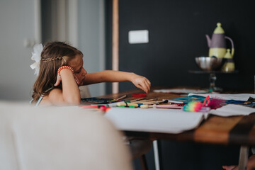 A young girl is focused on a creative art project at a table. Colorful art supplies like brushes and markers surround her, creating a vibrant atmosphere.