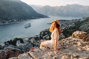 A woman enjoying a stunning view of Kotor Bay in Montenegro during sunset while sitting on a rocky...
