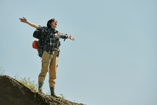 Young excited female backpacker keeping arms outstretched and shouting while standing on peak of mountain against blue sky