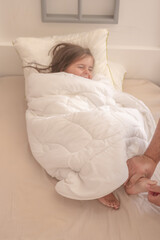 Little girl wrapped in a white blanket while sitting on the bed and smiling being tickled by dad