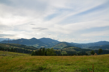 Lush green meadow, transitioning into rolling hills dotted with patches of trees and majestic mountain ranges stretch across the horizon under a partly cloudy sky. Carpathian Mountains, Ukraine