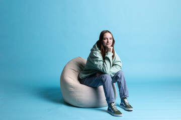 Portrait of happy girl sitting on beanbag armchair and smiling, isolated over blue studio background. Upbeat teenager seated on cozy bean bag, resting at home, having positive mood, studio backdrop