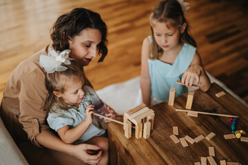 A mother and her daughters engage in creative play with wooden blocks. The family is bonding, exploring structure and imagination in a cozy home setting, emphasizing warmth and connection.