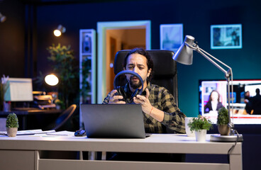 Male university student taking his wireless headset from table for music while using his laptop. Bearded man holding his headphones while seated in front of his portable computer.