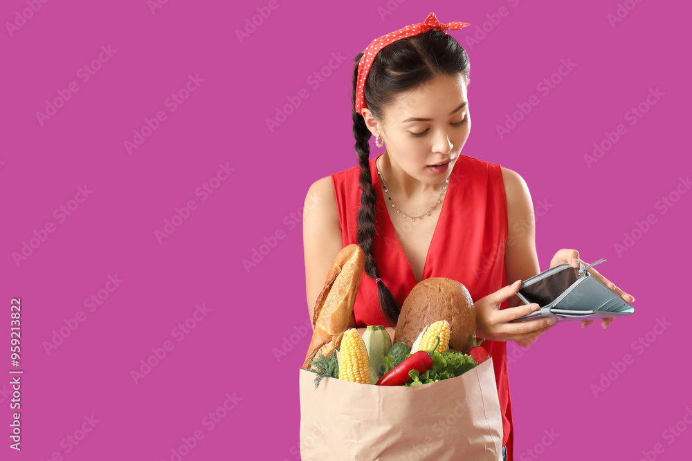 Wall mural shocked young asian woman holding paper bag with fresh products and wallet on purple background