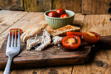 Plate with rustic food, fork, and knife on a wooden board. The plate contains chicken, bread, and tomatoes.