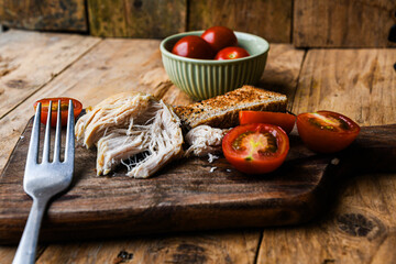 Plate with rustic food, fork, and knife on a wooden board. The plate contains chicken, bread, and tomatoes.