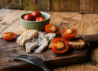 Plate with rustic food, fork, and knife on a wooden board. The plate contains chicken, bread, and tomatoes.