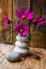 A vase with purple flowers sits on a wooden table beside a rock. The flowers are arranged in a way that they are all facing the same direction, creating a sense of harmony and balance