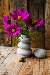 A vase with purple flowers sits on a wooden table beside a rock. The flowers are arranged in a way that they are all facing the same direction, creating a sense of harmony and balance